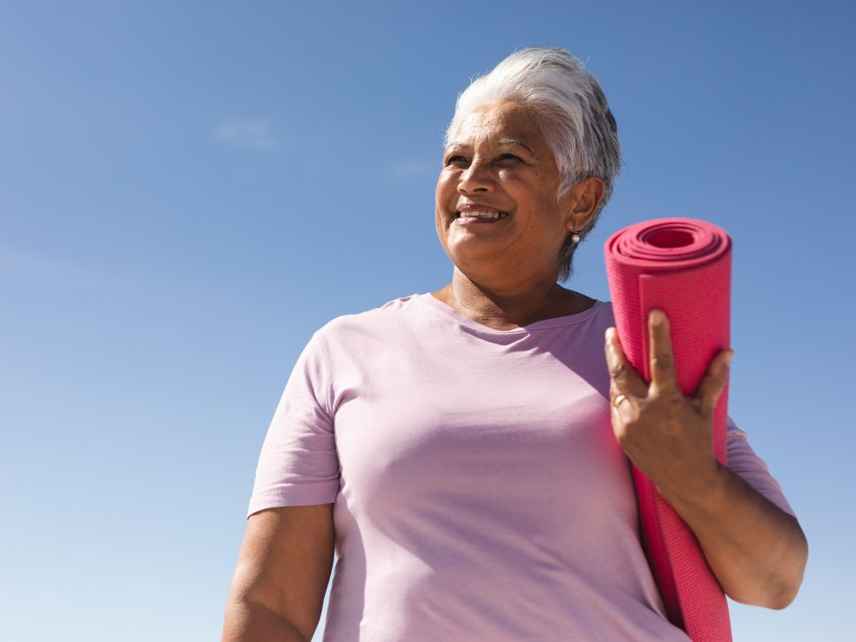 Smiling older woman holding a pink mat ready for a gentle yoga for seniors class